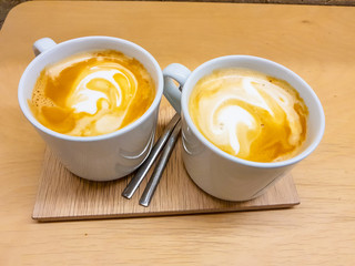 Two cups of latte with its foam drawing shapes. Presented on a wooden board in precious warm tones