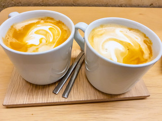 Two cups of latte with its foam drawing shapes. Presented on a wooden board in precious warm tones