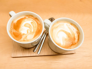 Two cups of latte with its foam drawing shapes. Presented on a wooden board in precious warm tones