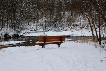 A winter scene of a park bench in the snow near the creek.
