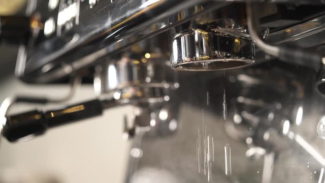 A close-up of coffee machine, drops of hot water fall down, then a hand fixes a portafilter, puts a cup underneath and the coffee starts pouring into it. Coffee making in a bar