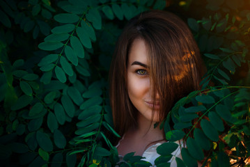 Beautiful girl with green eyes between green branches of trees. The charming look of the beautiful girl's eyes. Hair closes half of the face.