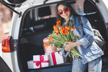 Young attractive woman in the car with presents box, gift and flowers. Beautiful lady in spring time with bouquet of tulips. Female in the automobile. © olenachukhil