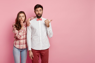 Horizontal view of shocked impressed female and male models say wow, notice something incredible at blank space, dressed in fashionable outfit, isolated over pink wall with free space for your text