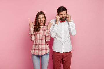 Joyful Caucasian woman makes horn gesture, exclaims as hears loud heavy metal, displeased bearded man plugs ears, doesnt like such music, have different tastes, isolated over pink background.
