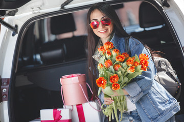 Young attractive woman in the car with presents box, gift and flowers. Beautiful lady in spring time with bouquet of tulips. Female in the automobile. © olenachukhil