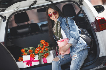 Young attractive woman in the car with presents box, gift and flowers. Beautiful lady in spring time with bouquet of tulips. Female in the automobile. © olenachukhil