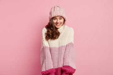 Horizontal shot of good looking woman with curly hair, looks with shy expression, wears warm hat and oversized knitted sweater, feels joyful, enjoys winter time, isolated over pink background.