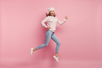 Horizontal shot of happy young Caucasian woman being photographed in motion, rejoices success, expresses positiveness, wears hat, jumper, jeans and sportshoes, jump high against pink background