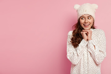 European beautiful girl keeps hands under chin, smiles broadly, had great weekend, wears childish headgear, stylish polka dot shirt, isolated over pink background with blank space aside for your text