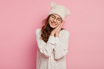 Happy young woman pretends sleeping, leans on hands, has tender smile, takes nap, wears transparent glasses, funny headgear with ears, stylish blouse, isolated over pink background, has rest