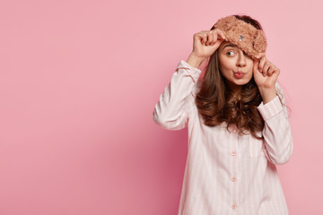 Indoor shot of funny young European woman wears eyemask, pyjamas, looks away, poses against pink...