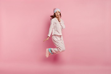 Portrait of surprised young lady wears casual pyjamas, eyemask and warm home boots, jumps in air,...