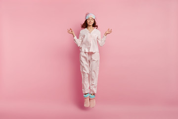 Relaxed beautiful woman meditates before having sleep, dressed in cotton pyjamas, warm chuni, eyemask, jumps against pink background, enjoys calm domestic atmosphere. Restful girl in nightsuit