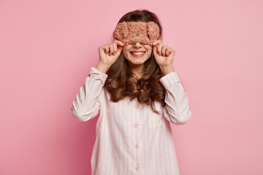 Photo cheerful dark haired woman with curly hair, covers eyes with warm eyemask, wears casual pyjamas, smiles gently, waits for surprise from husband in morning, isolated over pink background
