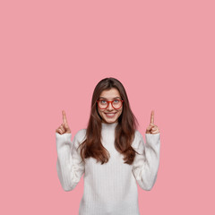Happy female seller points with both index fingers above, advertises new item, wears spectacles and casual white sweater, models against pink background, satisfied with discounts and special offers