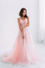 Luxurious brunette girl, with a bob haircut, posing in an expensive, pink dress with an open back. The princess in the white room. Scandinavian minimalist style interior. Image for prom and party