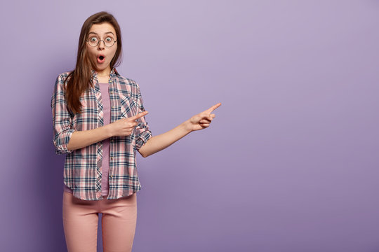 Studio shot of astonished woman with appealing appearance, shocked expression, dressed in checkered shirt, pink trousers, points at blank space, open mouth from surprise, isolated over purple wall