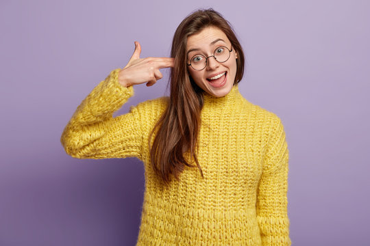 Photo of cheerful young European woman with dark hair, makes suicide gesture, dressed in yellow jumper, foolishes indoor against violet background. Human facial expressions and body language