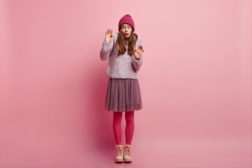 Horizontal shot of surprised frightened young lady keeps hands in protective gesture, afraids of phobia wears warm knitted hat, jumper full skirt, pink pantyhose and boots, models indoor. Fear concept