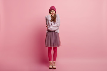 Studio shot of attractive pleasant looking woman feels overjoyed, comes on party with friends, dressed in stylish hat, oversized jumper, skirt, pink tights, models against studio background, laughs