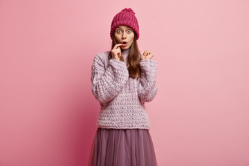 Puzzled scared hipster girl has bugged eyes, keeps fore finger on lips, wears winter hat, oversized long sleeved jumper, skirt, models over rosy background, has overwhelmed facial expression