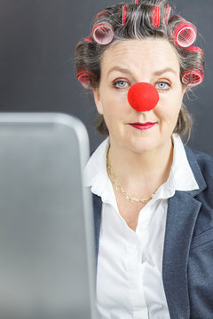 Frau tr&auml;gt eine rote Clownsnase und rote Lockenwickler und sitzt vor einem Laptop.