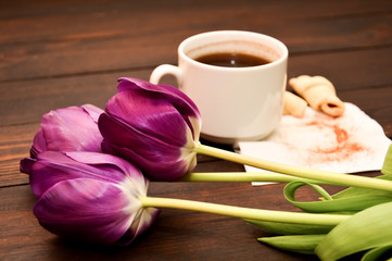 a cup of coffee or tea with a croissant on a napkin stands on a wooden background, next to it are purple tulips