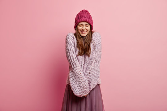Positive Caucasian young female model keeps hands together, wears long sleeved knitted sweater, pink hat, closes eyes from pleasure, models over rosy background. People and emotions concept.