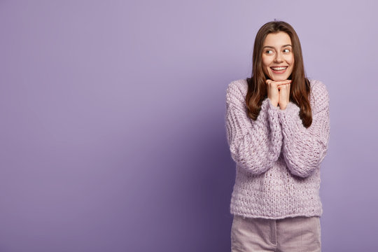 Image of pleased young lady has long dark hair, keeps hands together under chin, wears warm winter sweater, models over bright purple studio background, blank space for your advertising content
