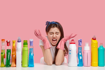 Indoor shot of dissatisfied cute housewife, spreads hands near face, sits at desktop with cleaning detergents, isolated over pink background, feels tired of working all day, cares of hygiene