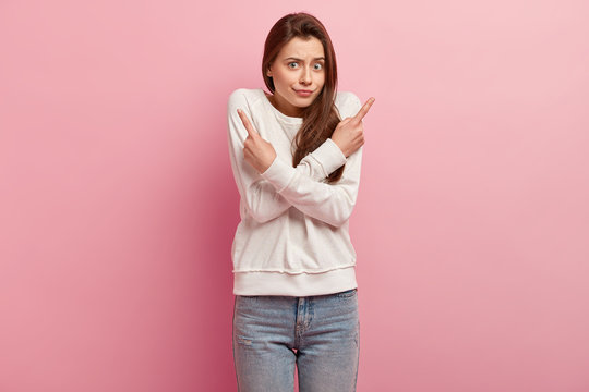Horizontal shot of puzzled lady crosses hands over chest, points with fore fingers, wears casual jumper and jeans, isolated over rosy background, has frustrated facial expression. People and choice