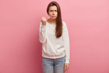 Threating concept. Angry woman raises clenched fist, has upset facial expression, promises to punish for bad behaviour, wears casual clothes, isolated on pink studio wall. Dont make me outraged