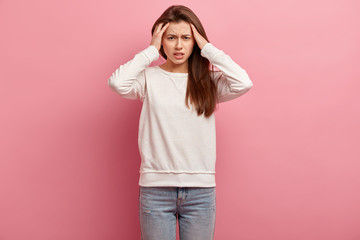 Pretty young European girl keeps hands on head, suffers from migraine, dressed in casual jumper and jeans, stands against pink background, feels under pressure, has headache, disappointment.