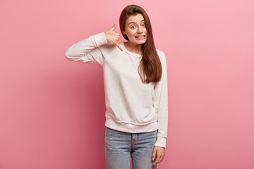 Caucasian beautiful young woman clenches teeth, makes phone gesture, wears sweatshirt and jeans, models against pink background, feels puzzled, keeps fingers like talking. Communication concept