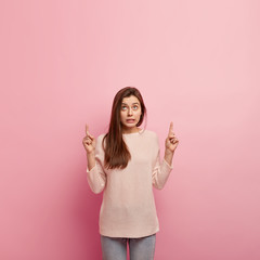 Vertical shot of puzzled European young woman points with both index fingers, advertises something...