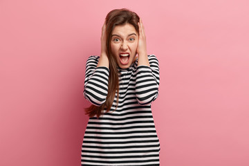 Headshot of frustrated young European lady keeps hands on ears, wears casual striped jumper, yells in displeasure, expresses negative emotions, isolated over pink background. Oh no, stop it please