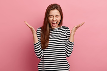Waist up shot of pleased young woman with dark hair, spreads palms, dressed in striped clothes, exclaims with happiness, reacts on something pleasant, cant believe in her success, poses indoor
