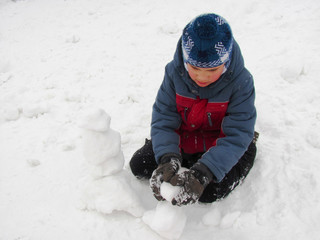 Fototapeta na wymiar Little boy in a winter warm clothes sits in the snow and makes a snowman