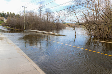 Water covering a flooded road. The yellow dividing line and sidewalk can be seen submerged. Trees...