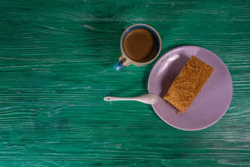 A piece of sponge cake and chocolates on a plate, next to it is a cup of coffee, photographed on a green wooden background close-up.