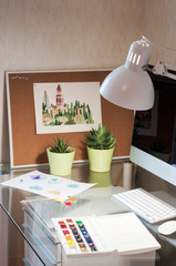 Glass desk with computer, succulents in green pots,  lamp, watercolor picture, watercolor paints and cork board. Modern desk workspace. Designer's workspace. 