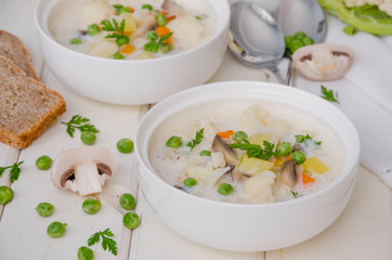 Cheese cream soup with cauliflower, mushrooms and green peas in a bowl on a white wooden background.