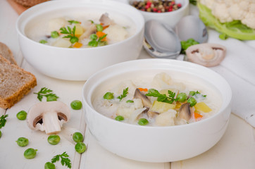 Cheese cream soup with cauliflower, mushrooms and green peas in a bowl on a white wooden background.