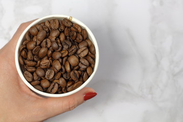 Coffee beans in a paper cup in female hande on a marble surface top view with copy space for text