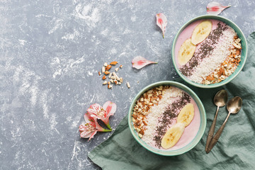 Yogurt with nuts, chia seeds, banana and coconut on a gray background, decorated with pink flowers. Healthy breakfast. Top view, copy space.