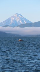 View of the Vilyuchinsky volcano from a tourist boat. The cloud lies on the coastal cliffs. Fishing boat is visible in the waters of Avacha Bay on the Kamchatka Peninsula, Russia.