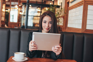 Beautiful charming brunette smiling Asian girl with tablet at table in cafe, student, freelancer or young manager