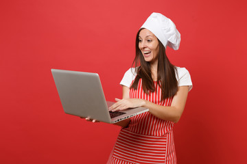 Housewife female chef cook or baker in striped apron, white t-shirt, toque chefs hat isolated on red wall background. Fun housekeeper woman looking for recipe in laptop pc. Mock up copy space concept.