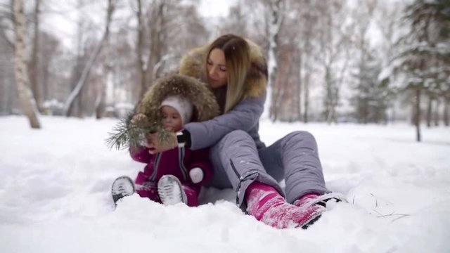 happy young mother with her cute little child are sitting on snow in yard outdoors, playing with branch
